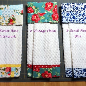 May include: Three white kitchen towels with different floral patterned fabric accents. The first towel has a patchwork design with a sweet rose pattern. The second towel has a vintage floral design with red roses on a teal background. The third towel has a scroll floral design with blue flowers on a white background. The text "1 Sweet Rose Patchwork", "2 Vintage Floral", and "3 Scroll Floral Blue" is printed below each towel.