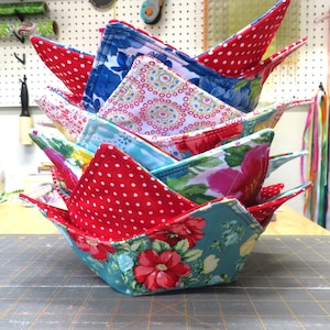 May include: A stack of colorful fabric bowl cozies with various patterns, including polka dots, floral designs, and geometric patterns. The cozies are made of cotton fabric and are designed to protect bowls from heat.