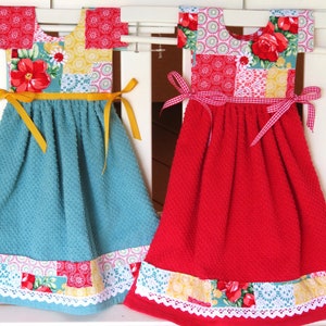 May include: Two decorative kitchen towels shaped like dresses with patchwork fabric and ribbon ties. The dress on the left is teal blue with yellow ribbon ties and the dress on the right is red with white and red gingham ribbon ties.