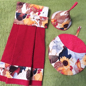 May include: A kitchen set featuring a red hand towel and two matching pot holders. The towel and pot holders have a decorative border with a rooster and sunflower pattern. The pot holders are round and have a red accent.