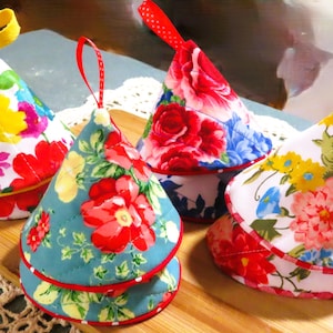 May include: Four colorful, cone-shaped fabric pot holders with floral patterns. Each pot holder features a different design with red, yellow, blue, and white flowers. They have a loop for hanging and are displayed on a wooden board.