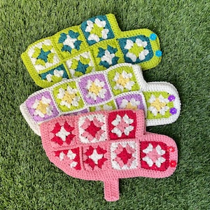 May include: Three handmade crochet leaf-shaped items in shades of green, purple, and pink. Each leaf features a patchwork design with floral squares in contrasting colors. The leaves are arranged on a bed of green grass.
