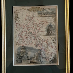 Op de afbeelding: Ingelijste antieke kaart van Oxfordshire, Engeland, met rode wegen en gedetailleerde illustraties van gebouwen en figuren. De kaart zit in een gouden lijst met een groene passe-partout.