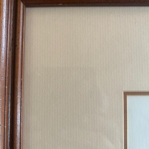 May include: A framed artwork with a brown wooden frame. The frame has a detailed, textured design. The artwork itself has a light beige background with a white inner border. The frame is a warm, natural wood tone.