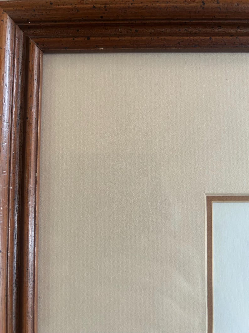 May include: A framed artwork with a brown wooden frame. The frame has a detailed, textured design. The artwork itself has a light beige background with a white inner border. The frame is a warm, natural wood tone.