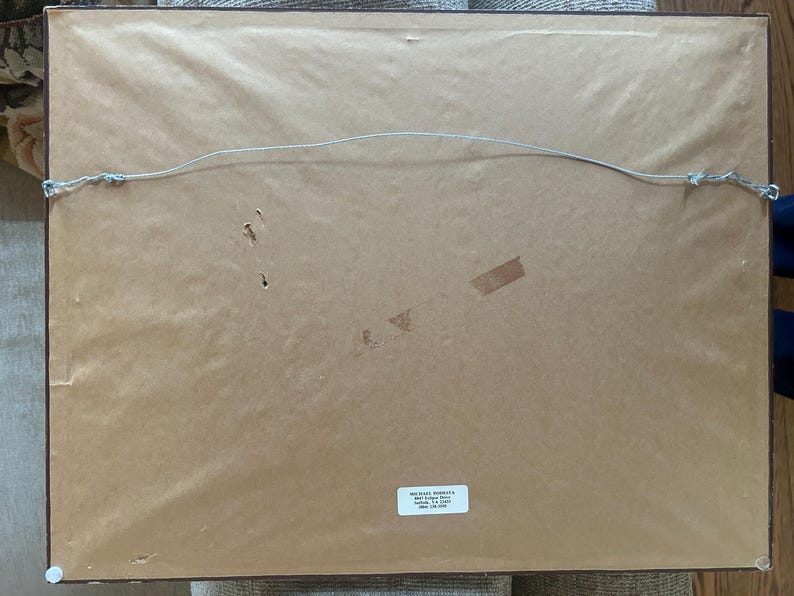 May include: Reverse side of a framed picture, featuring a brown paper backing, a hanging wire, and a small label with text. The frame is dark, and the paper shows signs of age and use.