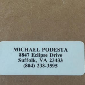 May include: A white rectangular label with rounded corners, featuring the name "MICHAEL PODESTA" in bold black font. Below, the address "8847 Eclipse Drive, Suffolk, VA 23433" and a phone number are printed.