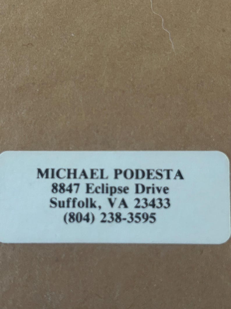 May include: A white rectangular label with rounded corners, featuring the name "MICHAEL PODESTA" in bold black font. Below, the address "8847 Eclipse Drive, Suffolk, VA 23433" and a phone number are printed.