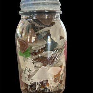 Op de afbeelding: Een glazen pot gevuld met diverse stukken zeeglas in tinten wit, bruin, groen en helder. De pot heeft een zilveren deksel en het woord "MASON" is in reliëf op de voorkant aangebracht. De pot staat tegen een zwarte achtergrond.