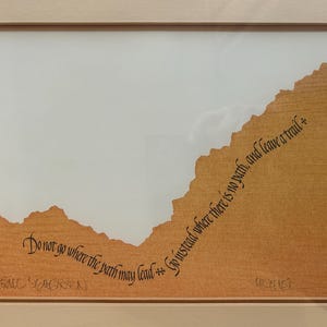May include: A framed artwork with a light brown textured background and a mountain-like shape. The artwork features the quote, "Do not go where the path may lead, go instead where there is no path, and leave a trail."
