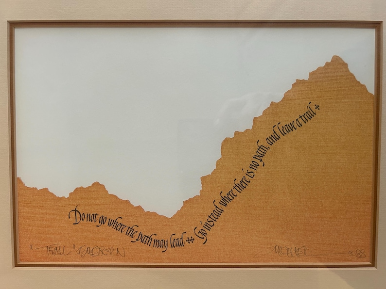 May include: A framed artwork with a light brown textured background and a mountain-like shape. The artwork features the quote, "Do not go where the path may lead, go instead where there is no path, and leave a trail."