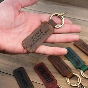 May include: A brown leather keychain with a gold ring, engraved with coordinates and the date "11/1/2019". Other keychains with various inscriptions and dates are visible.