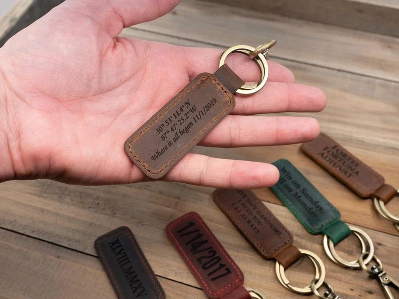 May include: A brown leather keychain with a gold ring, engraved with coordinates and the date "11/1/2019". Other keychains with various inscriptions and dates are visible.