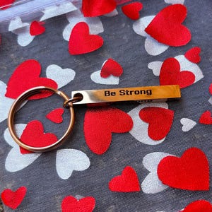 May include: A gold-coloured keychain with a ring and a bar that reads "Be Strong". The keychain is on a white background with red hearts.