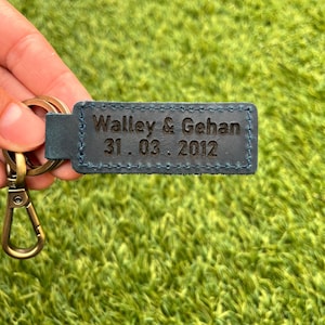 May include: A blue leather keychain with the engraved text "Walley & Gehan 31.03.2012". The keychain has a gold-colored clasp and ring. The background is a green, textured surface.
