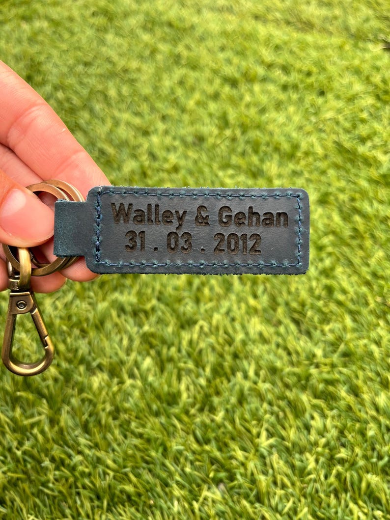 May include: A blue leather keychain with the engraved text "Walley & Gehan 31.03.2012". The keychain has a gold-colored clasp and ring. The background is a green, textured surface.