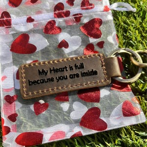 May include: A brown leather keychain with the engraved text "My Heart is full because you are inside" rests on a sheer bag printed with red and white hearts. The keychain has a gold-colored ring and is set against a backdrop of green grass.