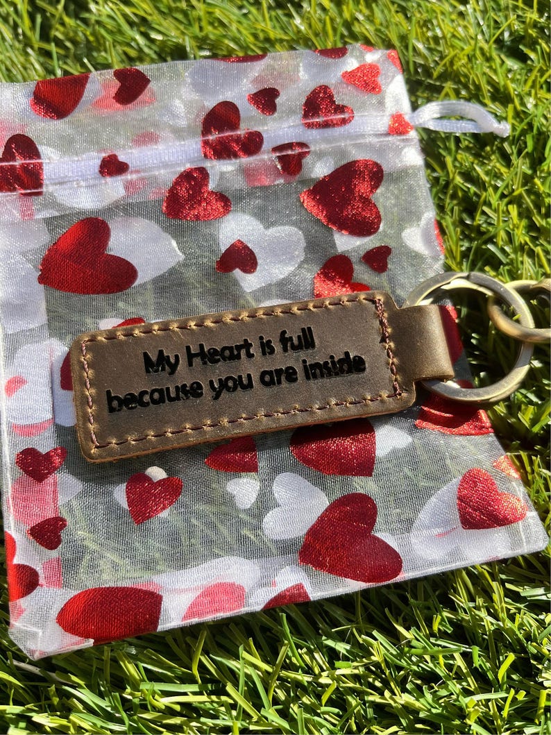 May include: A brown leather keychain with the engraved text "My Heart is full because you are inside" rests on a sheer bag printed with red and white hearts. The keychain has a gold-colored ring and is set against a backdrop of green grass.