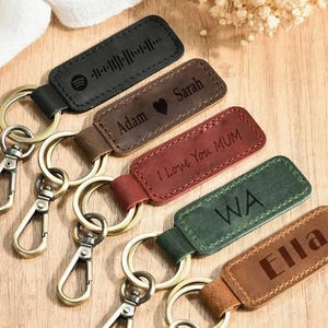 May include: A collection of personalized leather keychains in various colors, including black, brown, and red. Each keychain features a metal ring and clasp, with engraved text and designs. The keychains are arranged on a wooden surface.