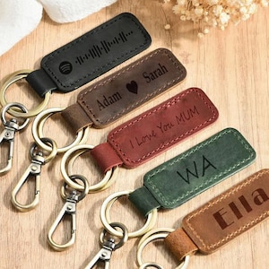May include: A collection of personalized leather keychains in various colors, including black, brown, and burgundy. Each keychain features a metal ring and clasp, with engraved text and designs. The keychains are arranged on a wooden surface.