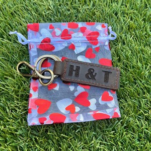 May include: A brown leather keychain with the embossed letters "H & T" rests on a sheer bag filled with red and white hearts. The keychain has a gold-colored metal ring and clasp. The bag is on a bed of green grass.