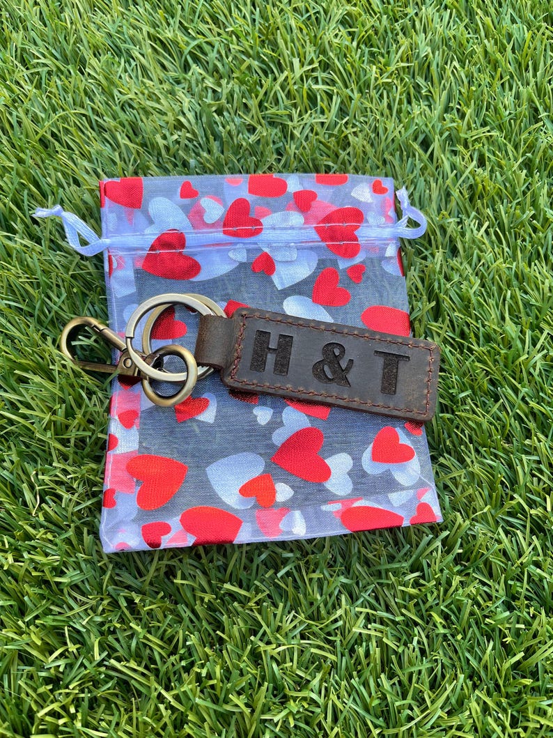 May include: A brown leather keychain with the embossed letters "H & T" rests on a sheer bag filled with red and white hearts. The keychain has a gold-colored metal ring and clasp. The bag is on a bed of green grass.
