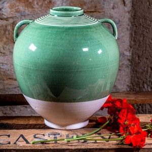Peut inclure: Un grand vase en céramique vert avec deux anses et une base blanche. Le vase est posé sur une caisse en bois avec le mot "CASTLE" imprimé dessus. Il y a des fleurs rouges devant le vase.