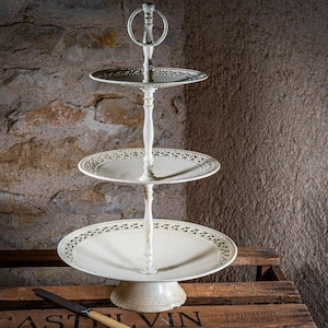 Könnte beinhalten: Ein weißer dreistöckiger Servierständer mit einem dekorativen, gewellten Rand an jedem Stockwerk. Der Ständer ist aus Metall und hat einen Vintage-Look.