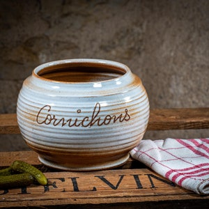 Può includere: Vaso di ceramica bianco con strisce marroni e la parola "Cornichons" incisa sul lato. Il vaso è appoggiato su una cassa di legno con un panno a quadri rosso e bianco e sottaceti verdi.