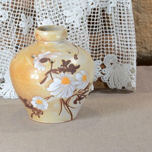Peut inclure: Un vase en céramique avec un corps arrondi et un col étroit. Le vase est peint avec une couleur de base jaune-beige et présente des marguerites blanches avec des centres jaunes et des accents bruns. L'arrière-plan comprend une nappe en dentelle blanche.