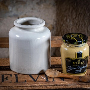 Op de afbeelding: Een stilleven met een witte keramische pot en een pot Dijon mosterd met een houten lepel. De mosterdpot heeft een zwart deksel met gouden letters en een etiket met de tekst "Maille Dijon Originale". De houten lepel ligt op een houten krat.