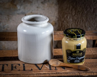 May include: A still life composition showing a white ceramic jar and a jar of Dijon mustard with a wooden spoon. The mustard jar has a black lid with gold lettering and a label that reads "Maille Dijon Originale". The wooden spoon rests on a wooden crate.