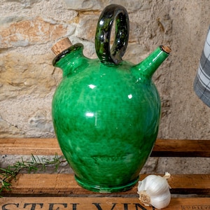 Könnte beinhalten: Eine grüne Keramik-Olivenölflasche mit einem Korkverschluss und einem Henkel. Die Flasche steht auf einer Holzkiste mit einer Knoblauchzehe und einem Rosmarinzweig.