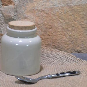 Può includere: Un piccolo barattolo di ceramica bianco sporco con coperchio in sughero, accanto a un cucchiaio d'argento. Il barattolo ha una forma arrotondata e una superficie liscia. Il cucchiaio ha un manico decorativo. Lo sfondo presenta un muro di pietra e tessuto di iuta.