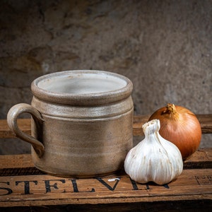 Può includere: Una tazza di ceramica marrone con manico si trova su una cassa di legno con una cipolla marrone e uno spicchio d'aglio bianco accanto.