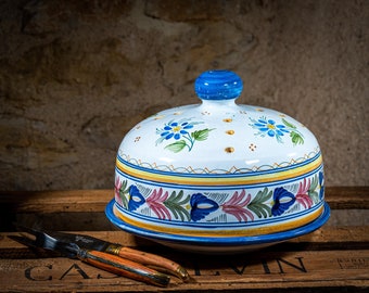Könnte beinhalten: Eine weiße Keramik-Glocke mit blauem und gelbem Blumendesign. Die Glocke hat einen gewölbten Deckel mit einem blauen Knopf und einer passenden Basis. Die Glocke steht auf einer Holzkiste.