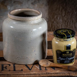 Pode incluir: Um pote de cerâmica branco com tampa marrom, um pote de mostarda Maille Dijon Original e uma colher de madeira em uma caixa de madeira. O pote tem a palavra "GLAMOY" impressa na parte inferior. O pote de mostarda tem uma tampa preta com letras douradas e o texto "Maille Dijon Originale" impresso no rótulo. A caixa tem a palavra "TELVIN" impressa nela.