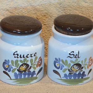 Op de afbeelding: Twee witte keramische potten met donkerbruine deksels. Op één pot staat het woord "Sucre" en op de andere "Sel" in zwart schrift. Beide potten zijn versierd met een bloemenmotief in blauw, geel en oranje.
