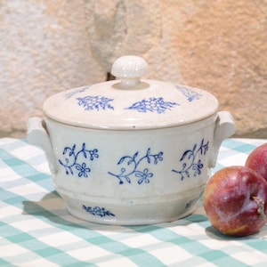 Peut inclure: Un plat de service en céramique blanche avec couvercle et motifs floraux bleus. Le plat a deux poignées et un petit bouton sur le couvercle. Deux prunes mûres sont posées à côté du plat sur une nappe à carreaux.