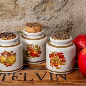 Pode incluir: Três potes de cerâmica com tampas de cortiça, cada um adornado com uma ilustração de fruta diferente. Os potes são brancos com detalhes marrons e estão sobre uma caixa de madeira. Maçãs e nozes estão ao lado dos potes.