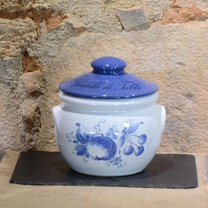Puede incluir: Un tarro de cerámica blanco con tapa y asas azules. El tarro está decorado con diseños azules de frutas y flores. La tapa tiene el texto "Poubelle de Table" en azul. El tarro está sobre una superficie negra.