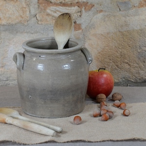 Puede incluir: Un jarrón rústico de gres gris con una cuchara de madera dentro. Una manzana roja, nueces y avellanas están dispuestas sobre una tela de arpillera. Dos cucharas de madera con cabezas de cepillo sugieren un tema de cocina.