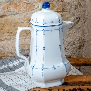 Könnte beinhalten: Eine weiße Porzellan-Kaffeekanne mit einem blauen und weißen geometrischen Muster. Die Kanne hat einen Deckel mit einem blauen Knopf und einen Ausguss an der Seite. Die Kanne steht auf einer Holzoberfläche mit einem weißen und grauen karierten Tuch.