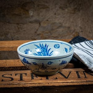 Op de afbeelding: Een witte keramische kom met blauwe bloemenmotieven. De kom staat op een houten kist met het woord "ASTREVIN" erop gedrukt.