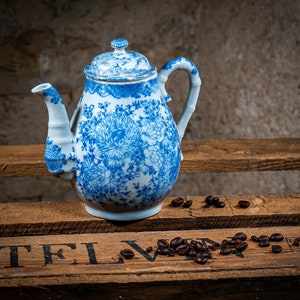 Peut inclure: Une théière en porcelaine bleue et blanche avec un motif floral et une anse en forme de bambou. La théière est posée sur une surface en bois avec des grains de café éparpillés autour. La théière a un couvercle assorti.