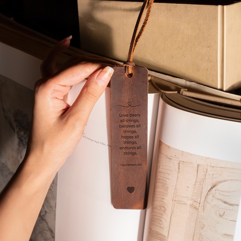 K&ouml;nnte beinhalten: Ein brauner Leder-Lesezeichen mit einer Lederschnur. Das Lesezeichen tr&auml;gt den Text "Love bears all things, believes all things, hopes all things, endures all things" und ein Herzsymbol. Das Lesezeichen befindet sich in einem Buch.