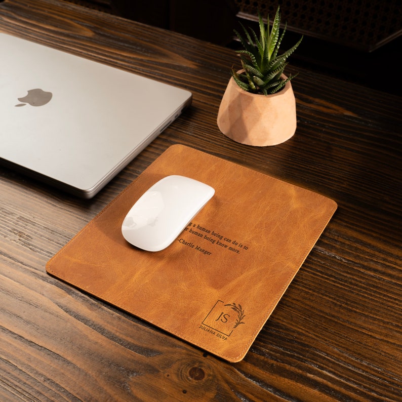 May include: A brown leather desk mat with a quote by Charlie Munger: "The most important thing a human being can do is to know more." The mat also has a monogram with the initials "IS" and the name "JULIANA SILVA" engraved on it.