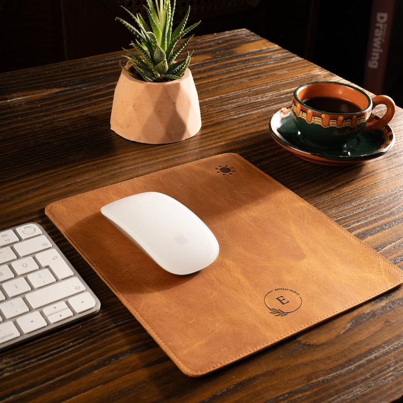 May include: A brown leather desk mat with a sun and a monogrammed initial "E" in a circle. A white wireless mouse is on the mat.