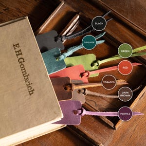 K&ouml;nnte beinhalten: Ein Buch mit dem Text "E.H. Gombrich" und mehrere Leder-Lesezeichen in verschiedenen Farben, darunter Schwarz, T&uuml;rkis, Gr&uuml;n, Rot, Tabak, Dunkelbraun und Lila. Die Lesezeichen sind in das Buch eingelegt und zeigen ihre unterschiedlichen Farbt&ouml;ne.