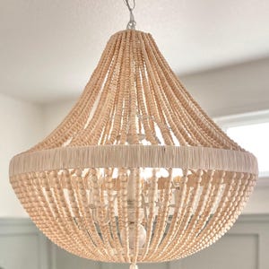 May include: A chandelier featuring a tiered frame draped with numerous strands of light beige wooden beads. The structure includes a central white metal fixture with multiple arms and a wide, circular band wrapped in light beige thread.